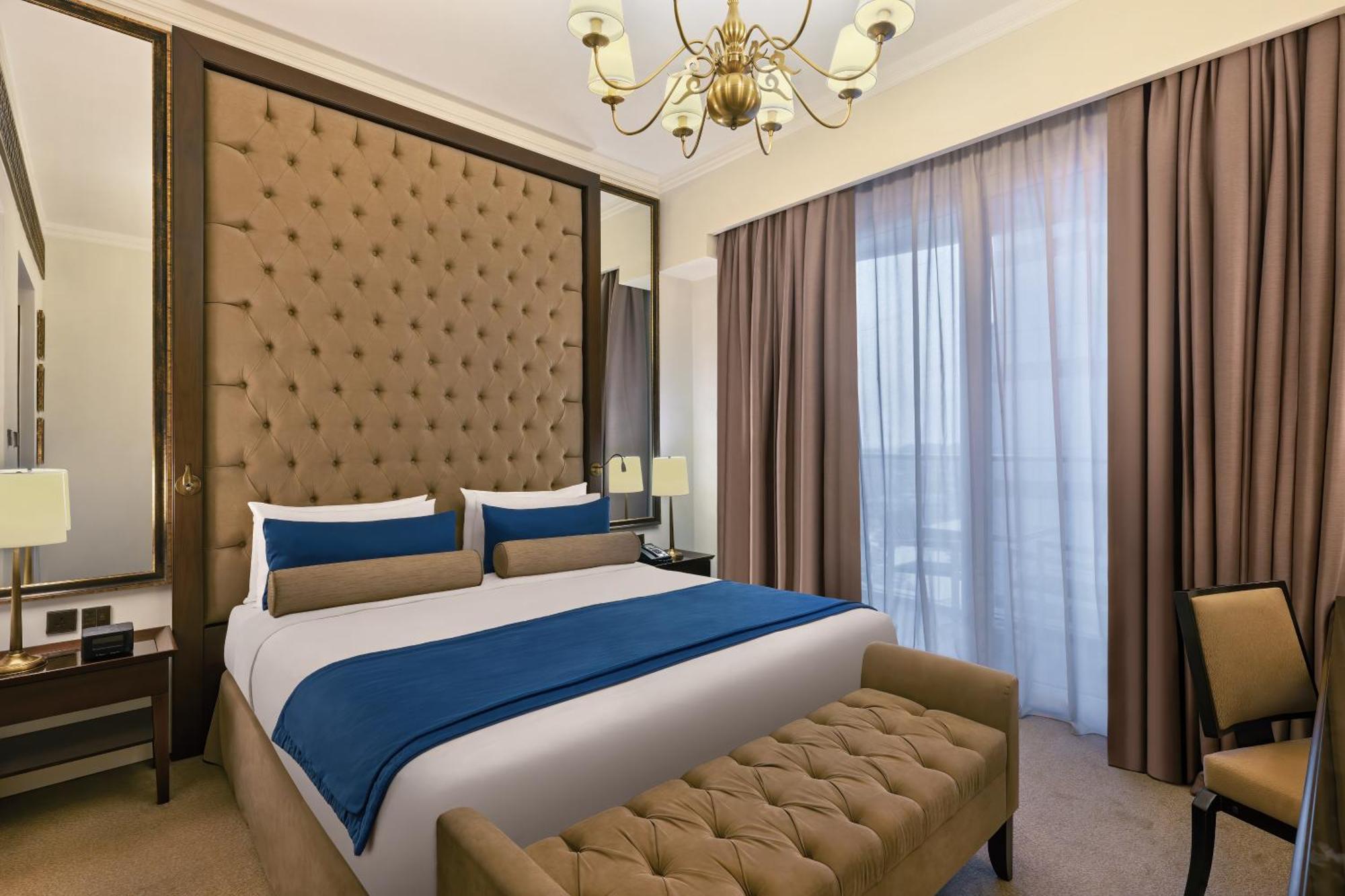דובאי Dukes The Palm, A Royal Hideaway Hotel מראה חיצוני תמונה The photo depicts a stylish hotel room with a modern design. It features a large bed with a tufted headboard upholstered in a tan color. The bed is dressed with white sheets and dark blue and beige decorative pillows. On either side of the bed, there